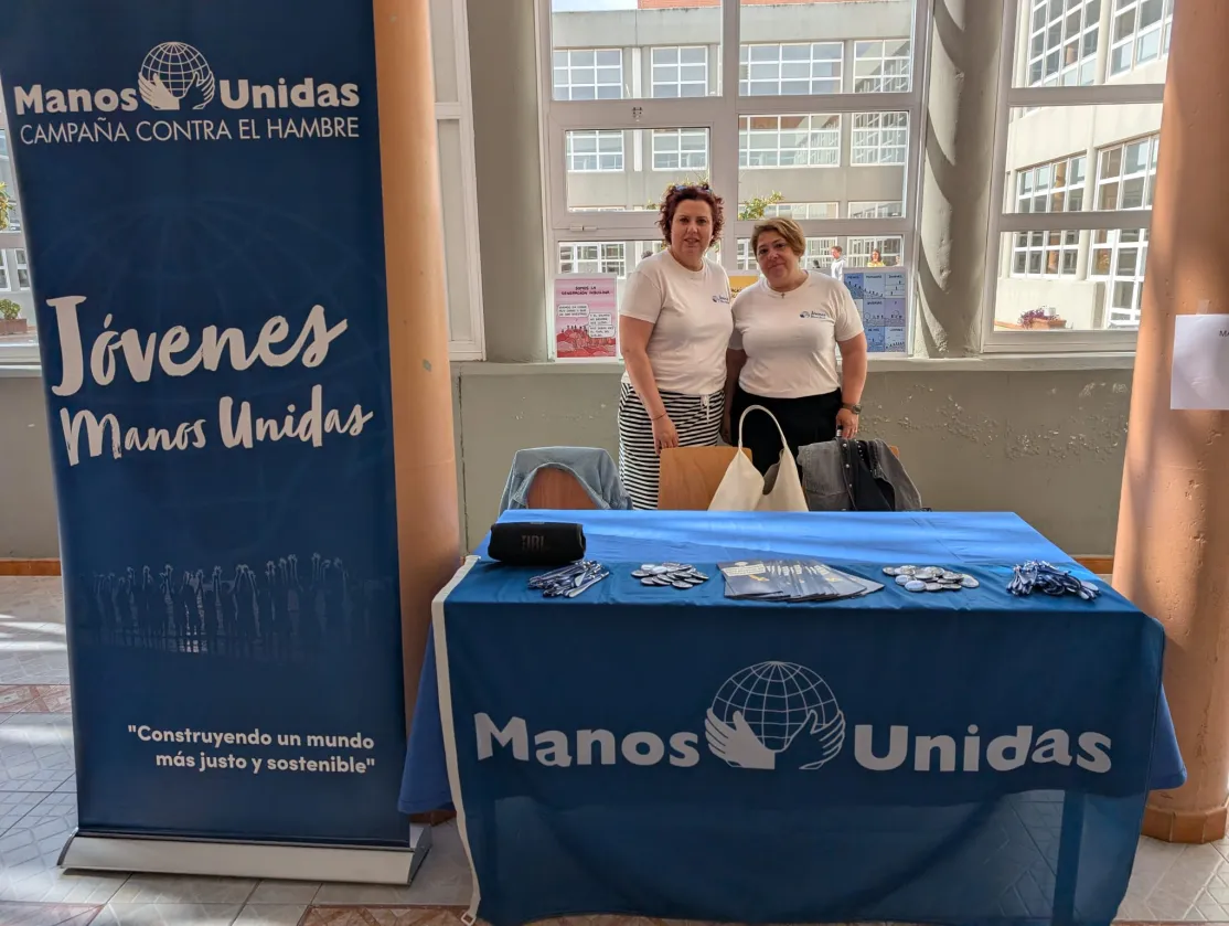 Dos representantes de Manos Unidas posan junto a un puesto informativo con material de la organización y un expositor dedicado al voluntariado joven.