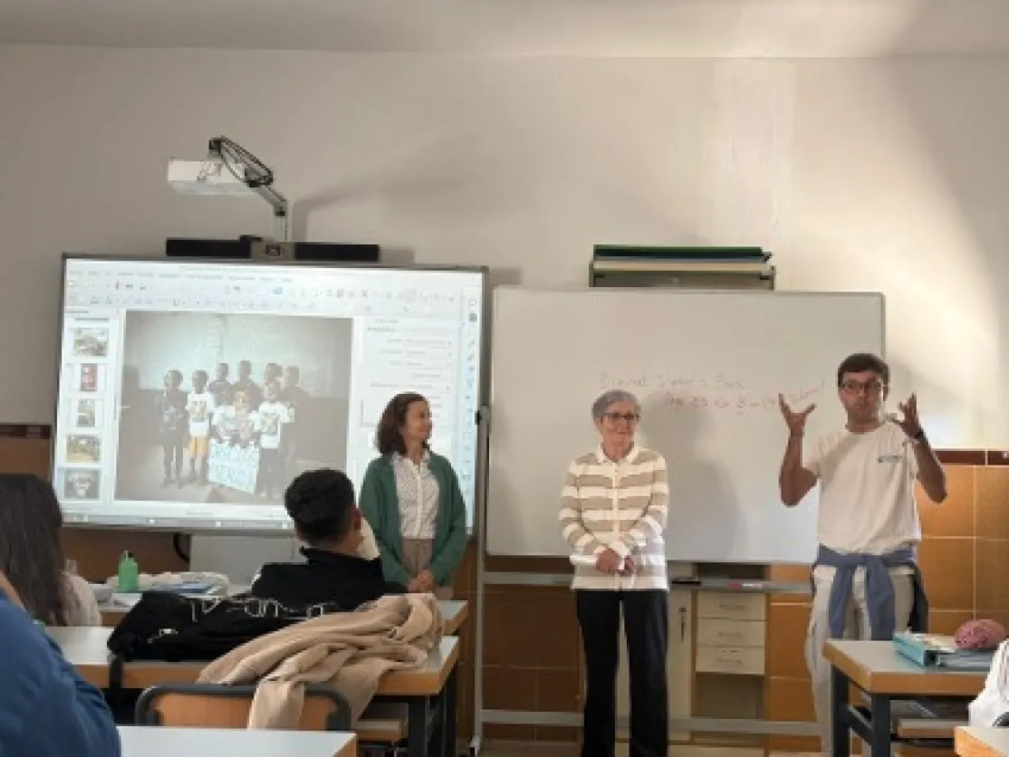 Tres integrantes de Manos Unidas realizan una presentación ante el alumnado en un aula, junto a una pantalla digital y una pizarra blanca.