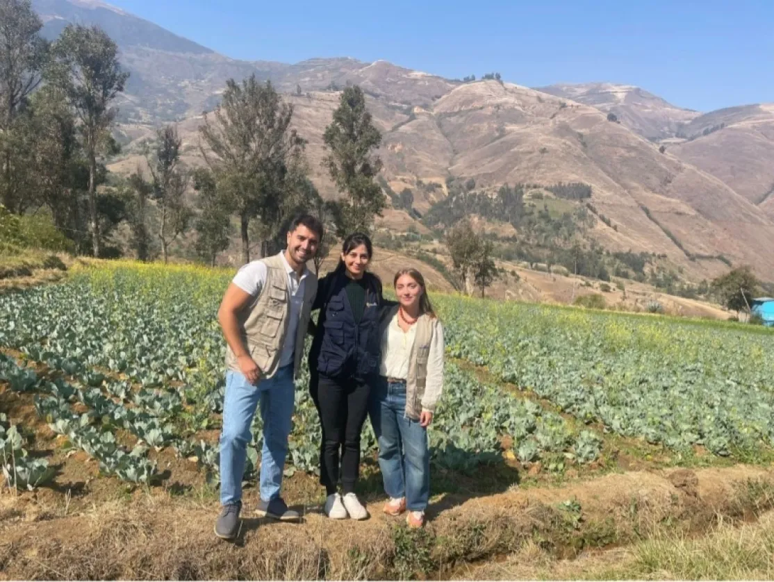 Tres personas posan sonrientes en un campo de cultivo, con árboles y montañas al fondo.