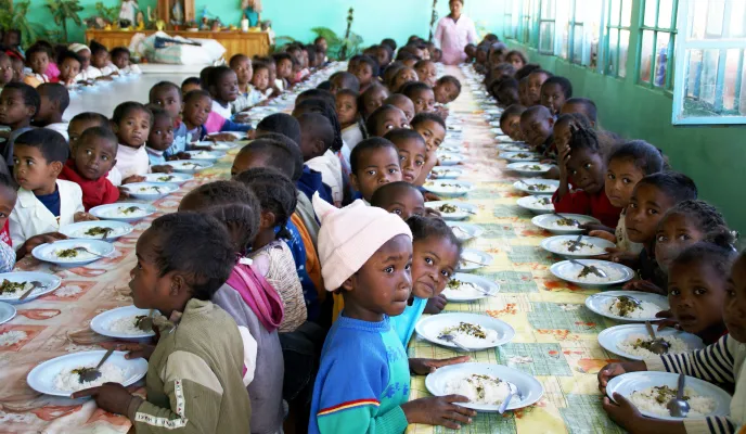 Numerosos niños están sentados alrededor de largas mesas mientras comen en un comedor escolar, acompañados por una mujer adulta al fondo.