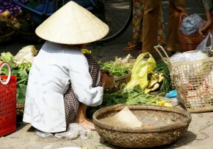 Una vendedora con sombrero cónico organiza verduras y productos frescos en un mercado al aire libre.
