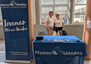 Dos representantes de Manos Unidas posan junto a un puesto informativo con material de la organización y un expositor dedicado al voluntariado joven.