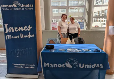 Dos representantes de Manos Unidas posan junto a un puesto informativo con material de la organización y un expositor dedicado al voluntariado joven.