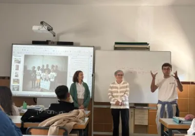 Tres integrantes de Manos Unidas realizan una presentación ante el alumnado en un aula, junto a una pantalla digital y una pizarra blanca.