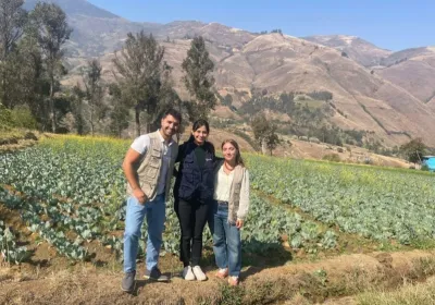 Tres personas posan sonrientes en un campo de cultivo, con árboles y montañas al fondo.