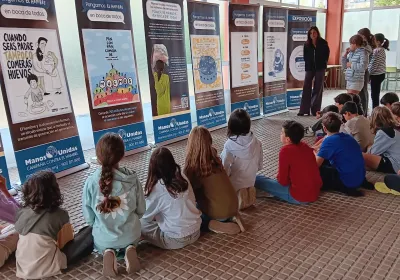 Un grupo de estudiantes sentado en el suelo escucha una explicación ante los paneles de la exposición «Los refranes de Manos Unidas» en un centro educativo.