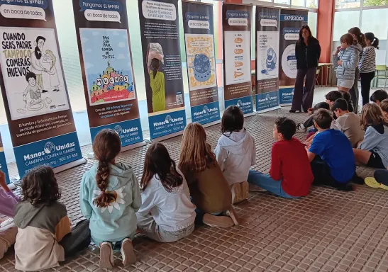 Un grupo de estudiantes sentado en el suelo escucha una explicación ante los paneles de la exposición «Los refranes de Manos Unidas» en un centro educativo.