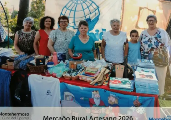 Voluntarias de Manos Unidas y una niña posan detrás del puesto solidario instalado en el Mercado Rural Artesano de Montehermoso, con productos artesanales, libros y materiales de la organización.