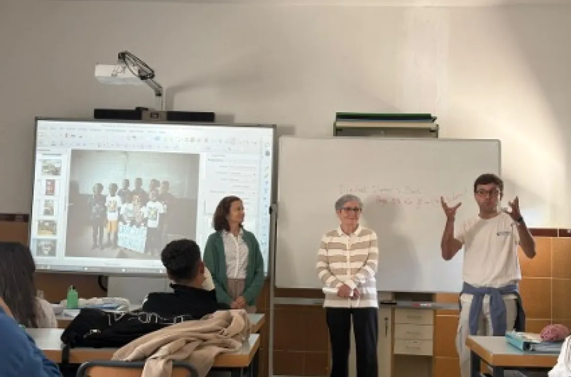 Tres integrantes de Manos Unidas realizan una presentación ante el alumnado en un aula, junto a una pantalla digital y una pizarra blanca.