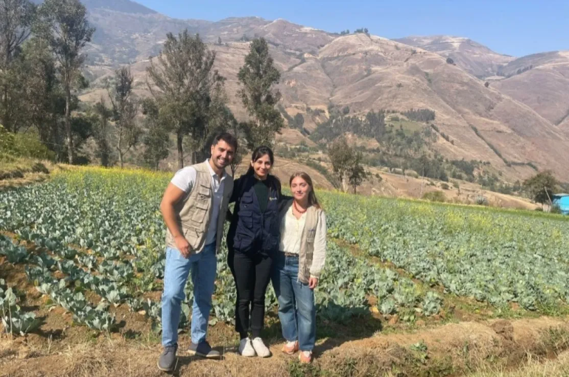 Tres personas posan sonrientes en un campo de cultivo, con árboles y montañas al fondo.