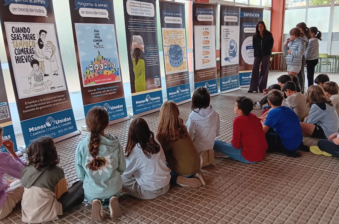 Un grupo de estudiantes sentado en el suelo escucha una explicación ante los paneles de la exposición «Los refranes de Manos Unidas» en un centro educativo.