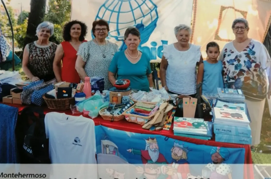Voluntarias de Manos Unidas y una niña posan detrás del puesto solidario instalado en el Mercado Rural Artesano de Montehermoso, con productos artesanales, libros y materiales de la organización.