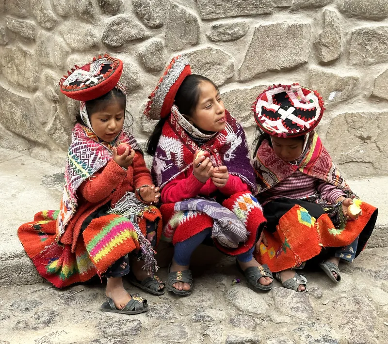 Tres niñas con vestimenta tradicional andina están sentadas junto a un muro de piedra mientras comen manzanas.