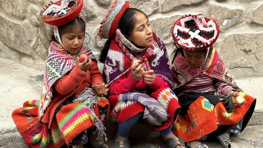 Tres niñas con vestimenta tradicional andina están sentadas junto a un muro de piedra mientras comen manzanas.