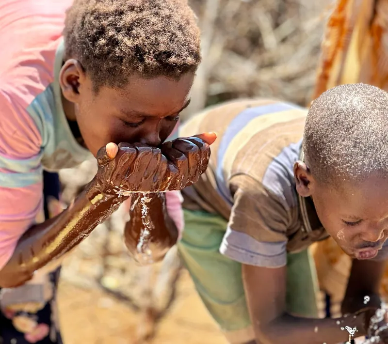 Dos niños beben agua directamente de sus manos junto a una fuente, rodeados por varias personas.