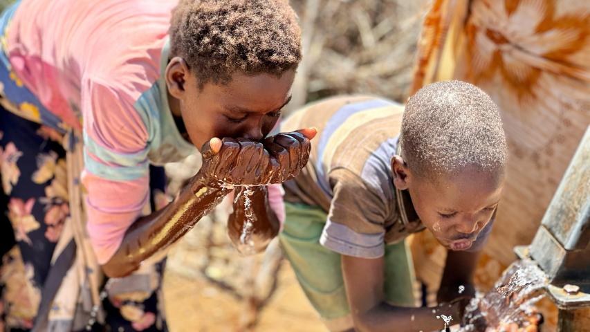 Dos niños beben agua directamente de sus manos junto a una fuente, rodeados por varias personas.