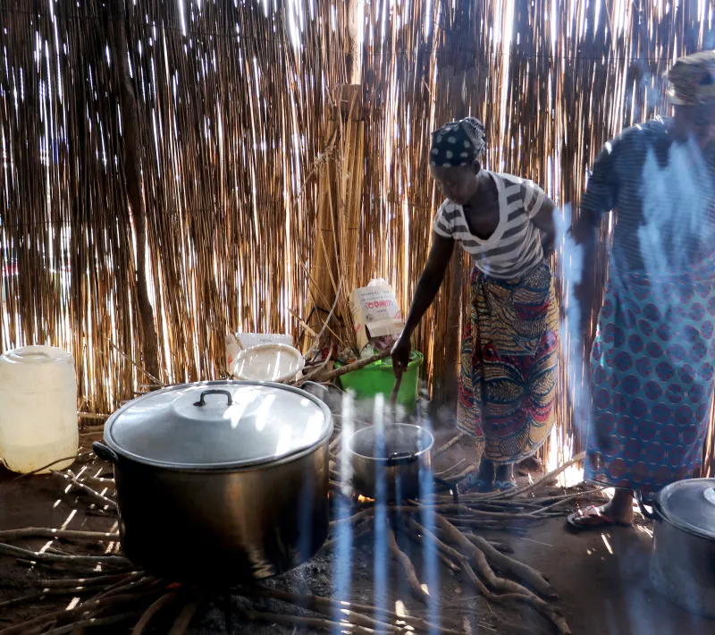 Dos mujeres cocinan en una estructura de cañas, rodeadas de grandes ollas, recipientes, leña y humo iluminado por la luz exterior.