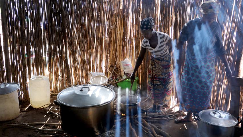 Dos mujeres cocinan en una estructura de cañas, rodeadas de grandes ollas, recipientes, leña y humo iluminado por la luz exterior.