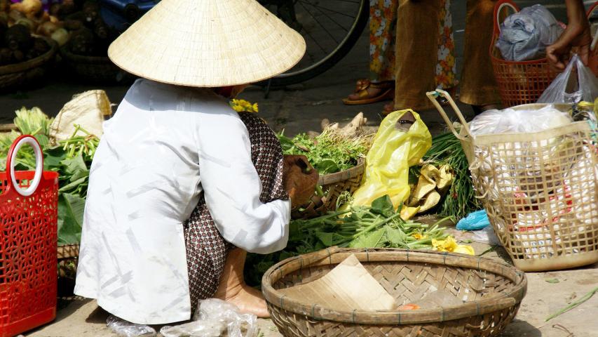 Una vendedora con sombrero cónico organiza verduras y productos frescos en un mercado al aire libre.