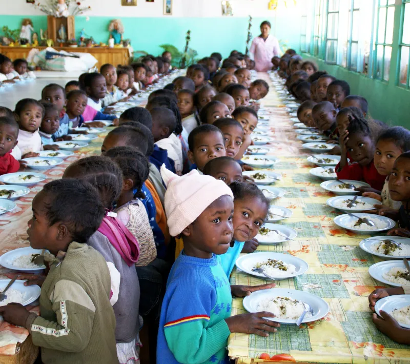 Numerosos niños están sentados alrededor de largas mesas mientras comen en un comedor escolar, acompañados por una mujer adulta al fondo.