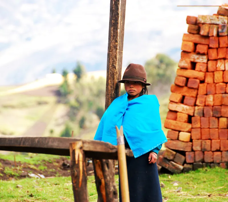 Una mujer con sombrero y poncho azul permanece de pie junto a una pila de ladrillos, con campos desenfocados al fondo.