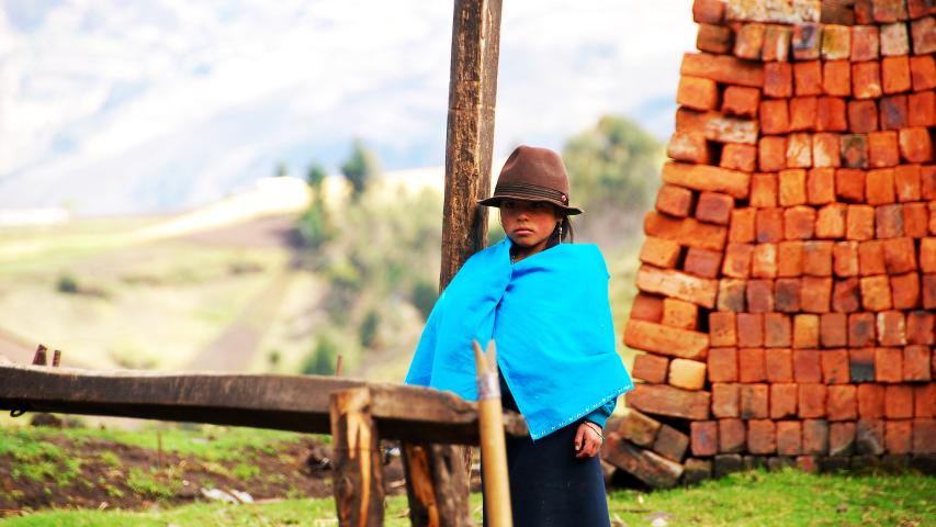 Una mujer con sombrero y poncho azul permanece de pie junto a una pila de ladrillos, con campos desenfocados al fondo.