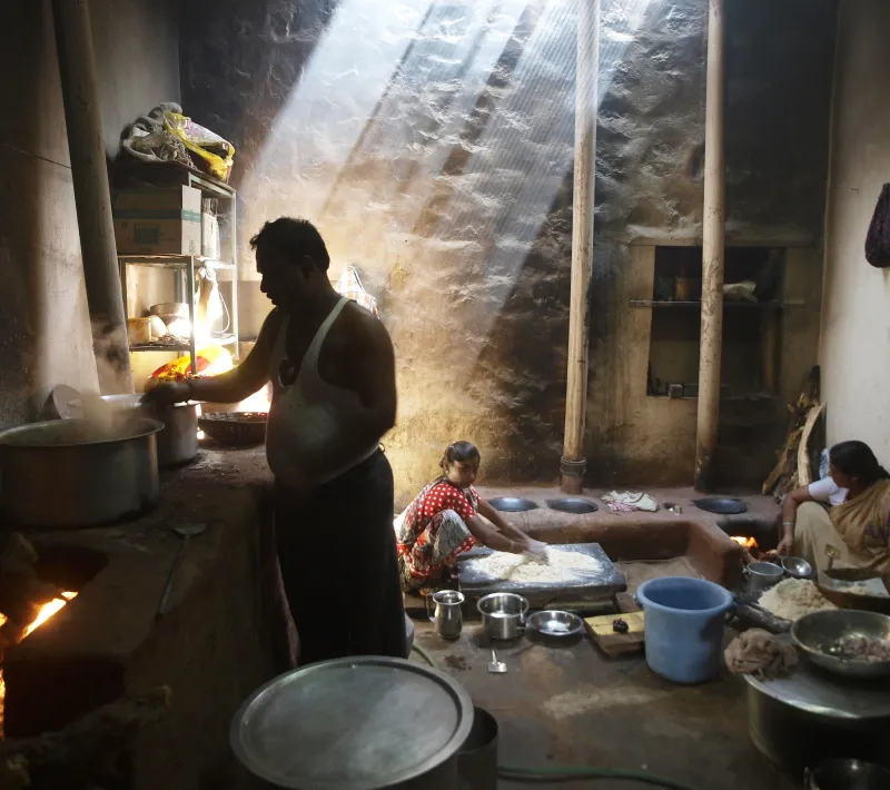 Varias personas preparan alimentos en una cocina tradicional con grandes ollas y fogones de leña, bajo haces de luz que entran desde el techo.