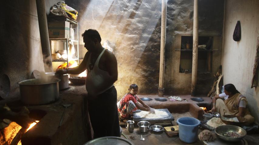 Varias personas preparan alimentos en una cocina tradicional con grandes ollas y fogones de leña, bajo haces de luz que entran desde el techo.