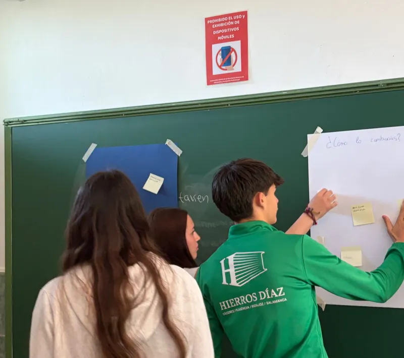 Tres estudiantes colocan notas adhesivas en un mural durante una actividad participativa realizada en el aula.
