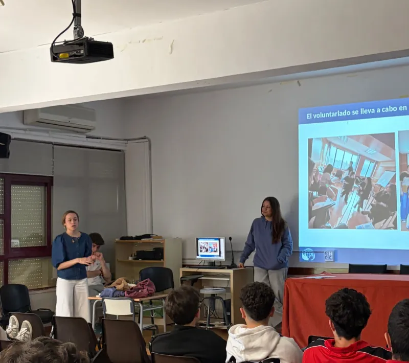 Dos jóvenes voluntarias de Manos Unidas imparten una charla ante el alumnado del IES El Brocense, con una presentación proyectada sobre el voluntariado.
