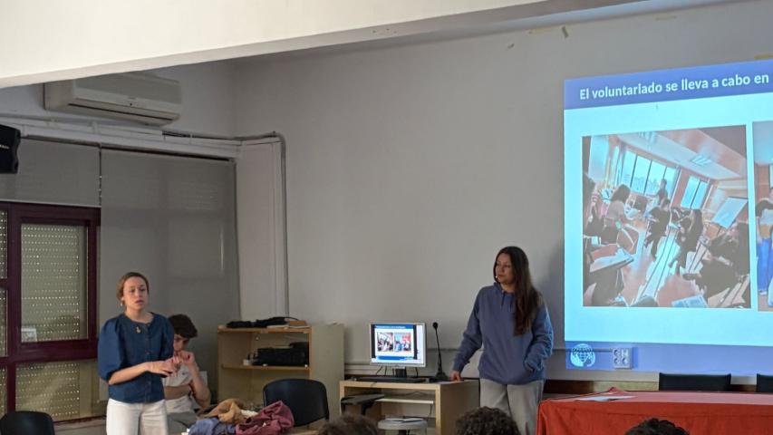 Dos jóvenes voluntarias de Manos Unidas imparten una charla ante el alumnado del IES El Brocense, con una presentación proyectada sobre el voluntariado.