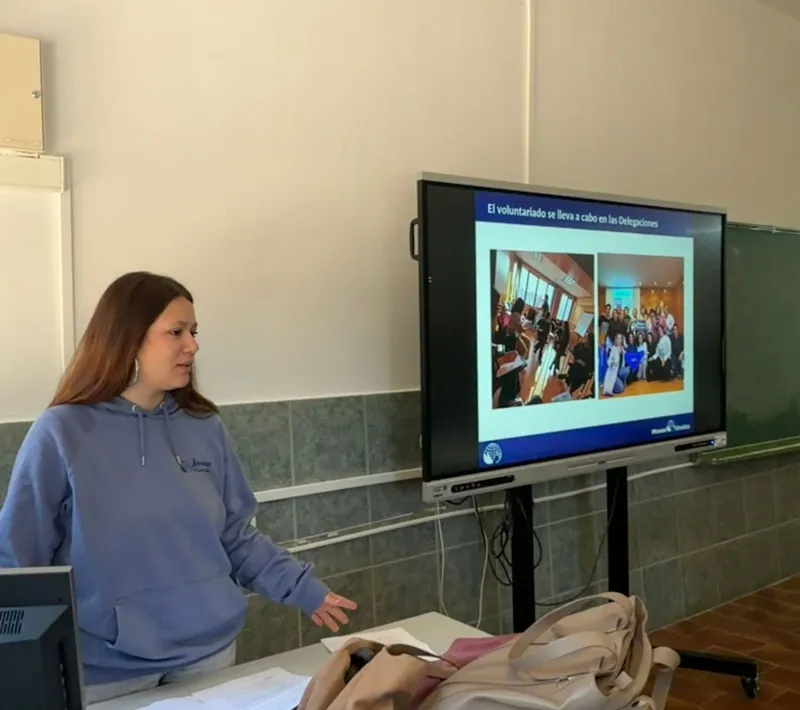 Una joven voluntaria presenta las actividades de voluntariado de Manos Unidas junto a una pantalla con imágenes de distintas acciones.