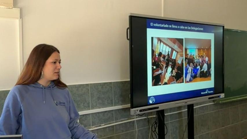 Una joven voluntaria presenta las actividades de voluntariado de Manos Unidas junto a una pantalla con imágenes de distintas acciones.