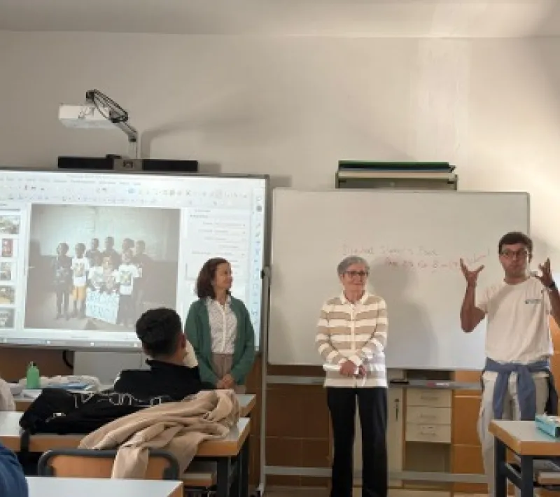 Tres integrantes de Manos Unidas realizan una presentación ante el alumnado en un aula, junto a una pantalla digital y una pizarra blanca.