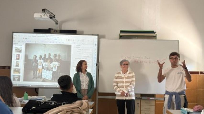 Tres integrantes de Manos Unidas realizan una presentación ante el alumnado en un aula, junto a una pantalla digital y una pizarra blanca.
