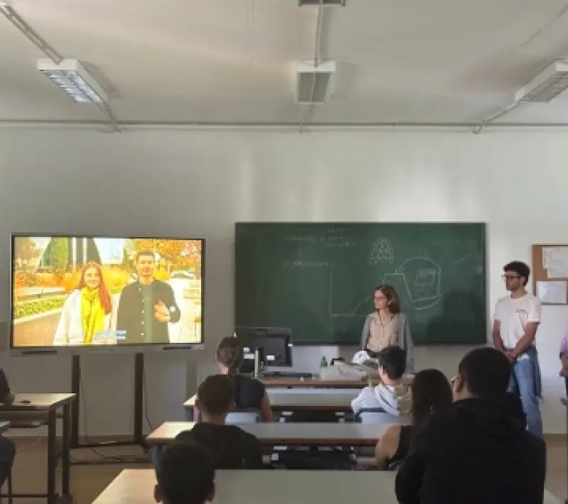 Miembros de Manos Unidas participan en una charla ante estudiantes, con un vídeo proyectado en una pantalla y contenidos escritos en la pizarra.