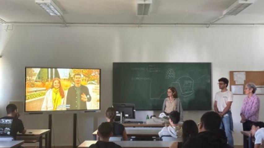Miembros de Manos Unidas participan en una charla ante estudiantes, con un vídeo proyectado en una pantalla y contenidos escritos en la pizarra.