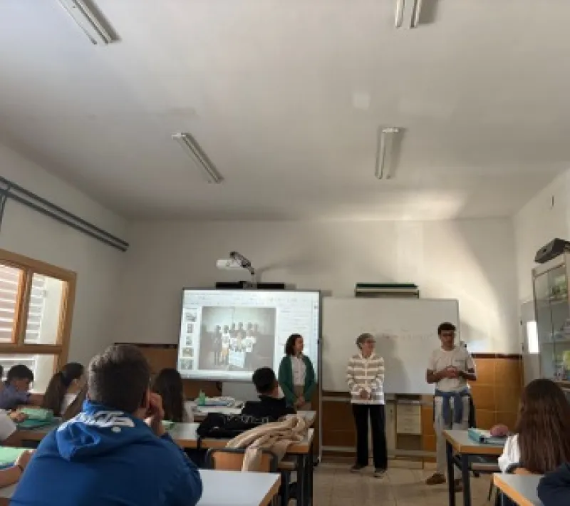 Vista general de un aula con estudiantes sentados mientras el equipo de Manos Unidas imparte una actividad educativa frente a una pantalla y una pizarra.