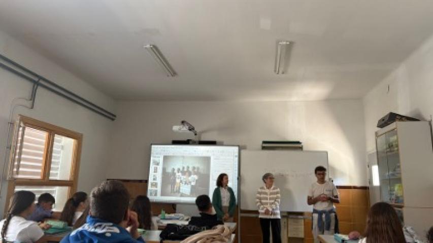 Vista general de un aula con estudiantes sentados mientras el equipo de Manos Unidas imparte una actividad educativa frente a una pantalla y una pizarra.