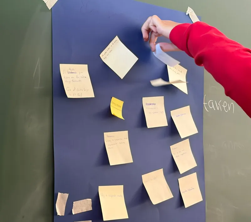 Primer plano de una mano pegando una nota adhesiva en un panel azul con distintas reflexiones escritas por el alumnado.