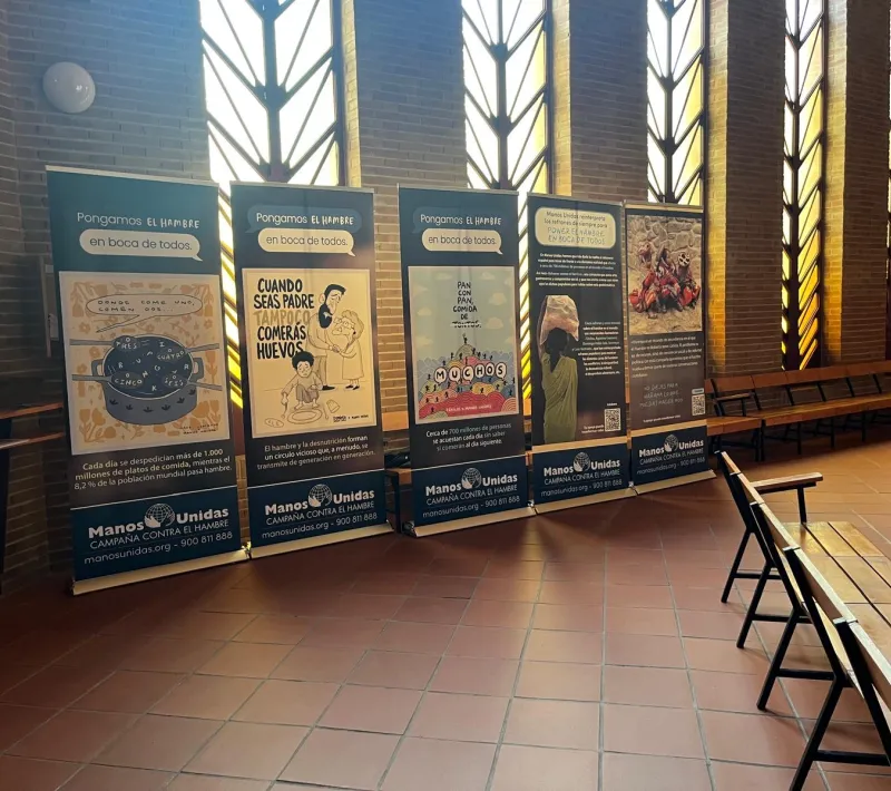 Cinco paneles de la exposición colocados junto a los bancos y las vidrieras del interior de una iglesia.