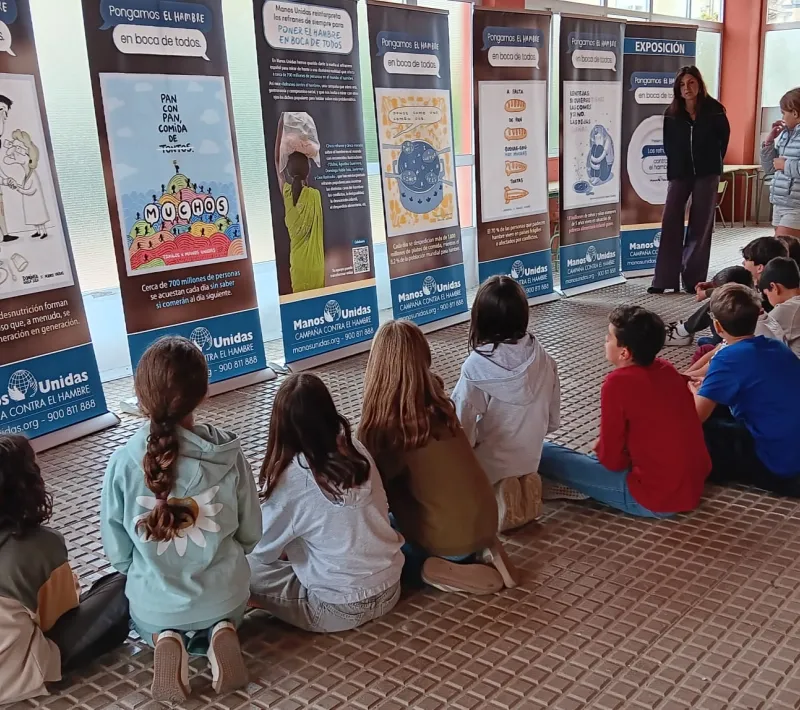 Un grupo de estudiantes sentado en el suelo escucha una explicación ante los paneles de la exposición «Los refranes de Manos Unidas» en un centro educativo.