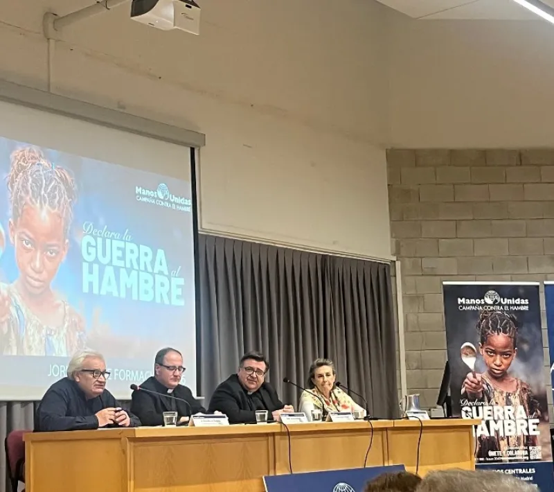 Cuatro ponentes intervienen en una mesa durante las Jornadas de Formación de Manos Unidas, junto a una pantalla y varios carteles de la campaña «Declara la guerra al hambre».